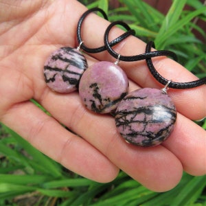 May include: Three pink and black round gemstone pendants on black cords. The pendants are made of polished stone with a natural pattern.
