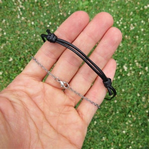 Pode incluir: Um colar de cord&atilde;o preto com uma corrente e um fecho prateados. O colar est&aacute; sendo segurado em uma m&atilde;o.