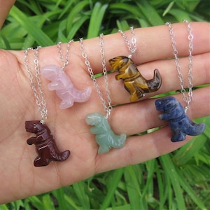 May include: Five silver necklaces with dinosaur-shaped pendants made of different gemstones. The pendants are in the colors pink, brown, green, blue, and tiger's eye.
