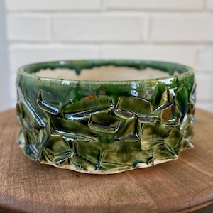 May include: A round ceramic bowl with a glossy, deep green glaze. The exterior features a textured, faceted surface with irregular, angular indentations that create a crinkled appearance. The interior of the bowl is smooth and off-white in color.