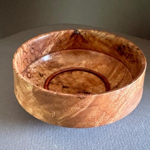 May include: A small, round wooden bowl with a natural wood finish. The bowl has a dark brown rim and a light brown interior. The wood grain is visible throughout the bowl.