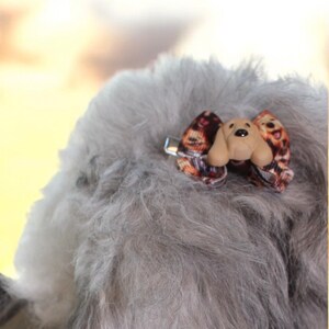 May include: A small dog bow with a tan dog face and ears, and a patterned bow. The bow is attached to a silver metal clip. The bow is attached to gray fur.