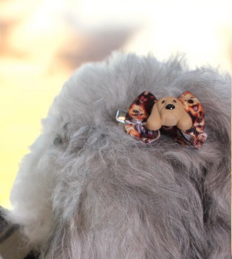 May include: A small dog bow with a tan dog face and ears, and a patterned bow. The bow is attached to a silver metal clip. The bow is attached to gray fur.