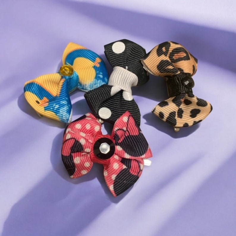 May include: A collection of small, decorative hair bows in various patterns. The bows feature designs such as polka dots, leopard print, and abstract patterns in colors like yellow, blue, black, and red. Each bow has a small clip for easy attachment.