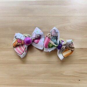 May include: Two white fabric hair bows with cupcake designs. Each bow features a colorful cupcake illustration with sprinkles and a decorative center. One bow has a purple center, and the other has a purple bow.