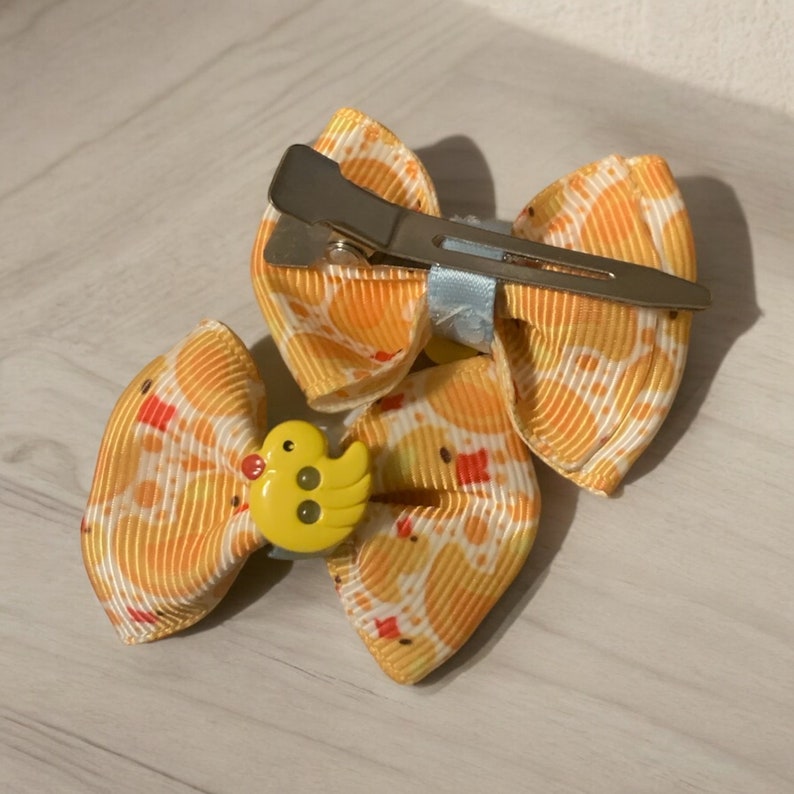 May include: A yellow and white patterned hair bow with a metal clip. The bow features a yellow duck embellishment in the center. The bow is made of a ribbon material.