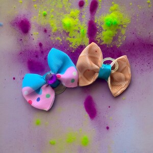 May include: Two colorful pet hair bows. One bow is light blue and pink with polka dots and a purple gem. The other bow is tan with a teal center. The background is a light purple with splashes of neon green and purple.
