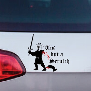 May include: Black vinyl car sticker depicting a cartoon knight with a sword and the words "'Tis but a scratch" in a gothic font. The sticker is on the rear of a silver car, near the rear light.