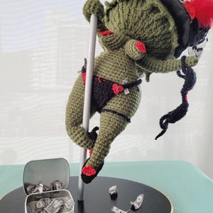 May include: A handmade, green crocheted figure in a black bikini, red accents, and a black hat with red feathers, posed on a silver pole. A small tin with dollar bills sits nearby, with more bills scattered around the base.