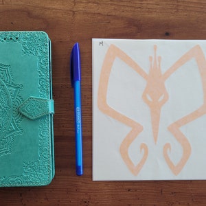 May include: A teal leather journal with an intricate mandala design and a decorative clasp. A blue pen and a white sticker with a peach-colored butterfly design are also pictured. The sticker has the letter "M" written on it.