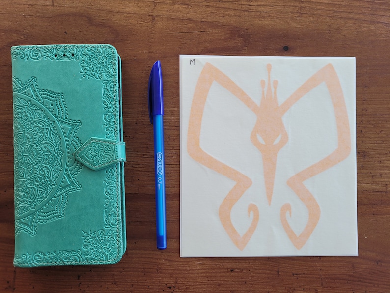 May include: A teal leather journal with an intricate mandala design and a decorative clasp. A blue pen and a white sticker with a peach-colored butterfly design are also pictured. The sticker has the letter "M" written on it.