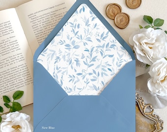 May include: A dusty blue envelope with a white interior liner featuring a blue botanical leaf pattern. The envelope has the text New Blue printed on the lower left corner. It is surrounded by an open book, white roses, and gold wax seals.