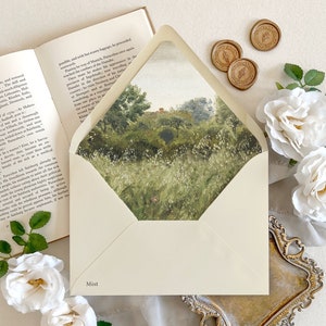 May include: A white envelope with a watercolor painting of a green landscape on the front. The envelope has the word "Mist" printed on the bottom. The envelope is surrounded by white roses, gold wax seals, and a book.