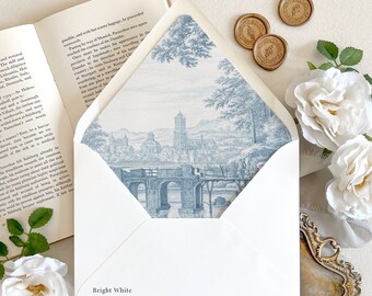 May include: A bright white envelope with a blue and white toile de jouy landscape print on the interior flap, featuring a bridge and cathedral scene. The envelope is displayed alongside an open book, three gold wax seals, and white roses.