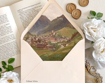 May include: A cream-colored envelope with a Vellum White label, featuring an interior liner printed with a detailed landscape painting of a mountain village. The envelope is displayed alongside an open book, three wax seals, and white roses.