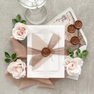 May include: A set of white and cream wedding invitations with a brown ribbon and wax seal. The invitations are decorated with delicate pink roses and greenery.
