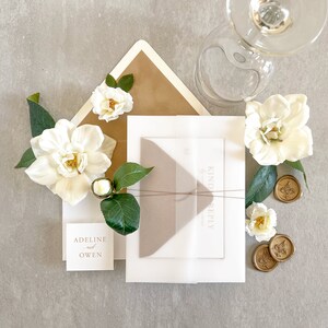 May include: A white envelope with a brown envelope inside. The envelopes are decorated with white flowers and gold wax seals. The text "Kindly Reply" is printed on the white envelope. The text "Adeline & Owen" is printed on a small white card.