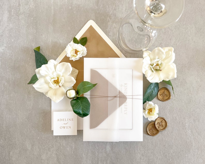 May include: A white envelope with a brown envelope inside. The envelopes are decorated with white flowers and gold wax seals. The text "Kindly Reply" is printed on the white envelope. The text "Adeline & Owen" is printed on a small white card.