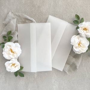 May include: Three white envelopes with a white fabric and white roses on a gray background.