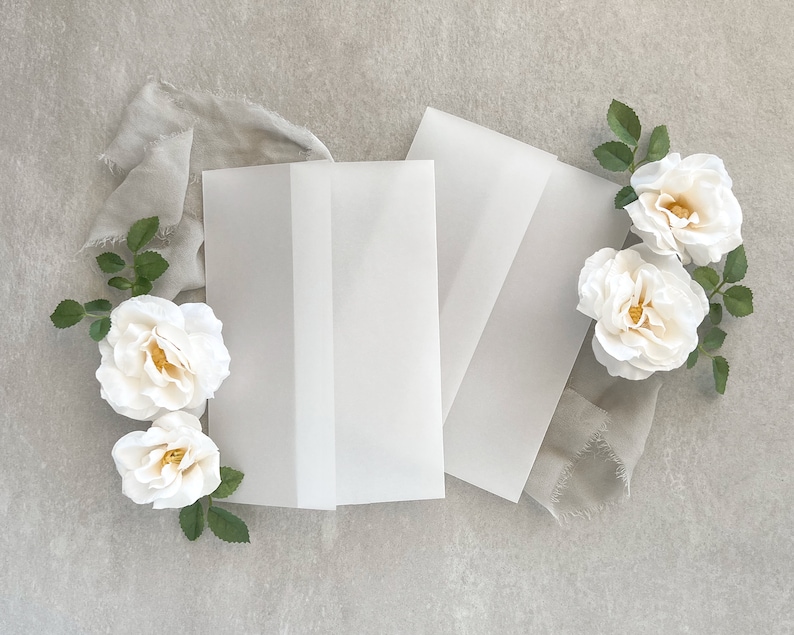 May include: Three white envelopes with a white fabric and white roses on a gray background.