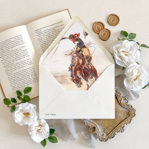 Op de afbeelding: Een zachtwitte envelop met een bedrukte voering met een cowboy op een bokkend paard die met een lasso zwaait. De envelop wordt getoond naast een open boek, drie gouden waszegels en verschillende witte rozen met groene bladeren.