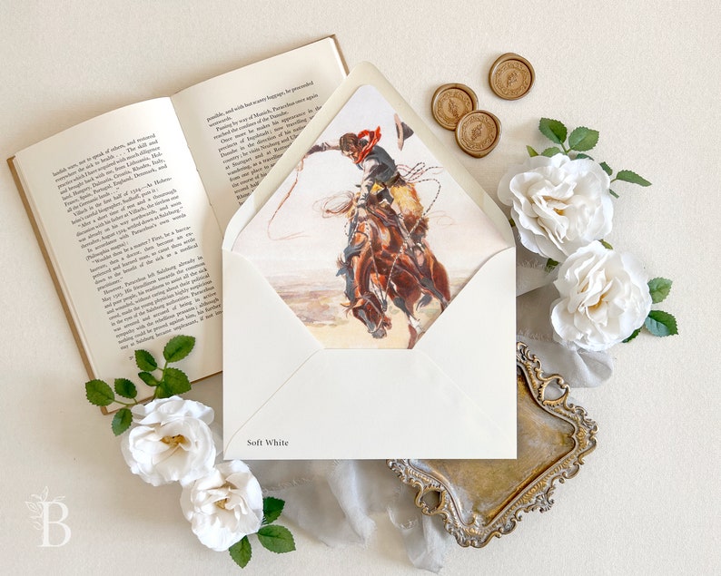 Op de afbeelding: Een zachtwitte envelop met een bedrukte voering met een cowboy op een bokkend paard die met een lasso zwaait. De envelop wordt getoond naast een open boek, drie gouden waszegels en verschillende witte rozen met groene bladeren.