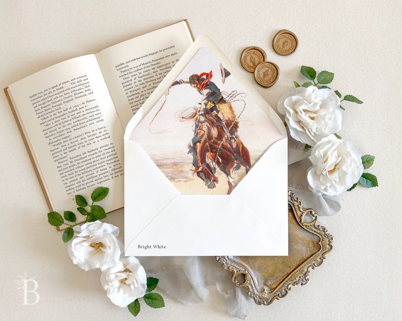 Op de afbeelding: Een helderwitte envelop met een gedrukte illustratie van een cowboy op een bokkend paard aan de binnenkant van de flap. De envelop wordt getoond naast een open boek, drie gouden waszegels en verschillende witte rozen met groene bladeren.