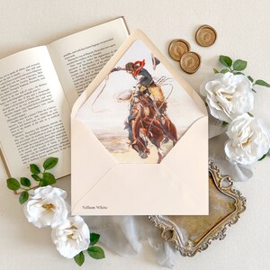 Op de afbeelding: Een cr&egrave;mekleurige envelop met een Vellum White label, voorzien van een binnenvoering met een illustratie van een cowboy op een bokkend paard met een lasso. De envelop ligt naast een open boek, drie gouden waszegels en witte rozen.