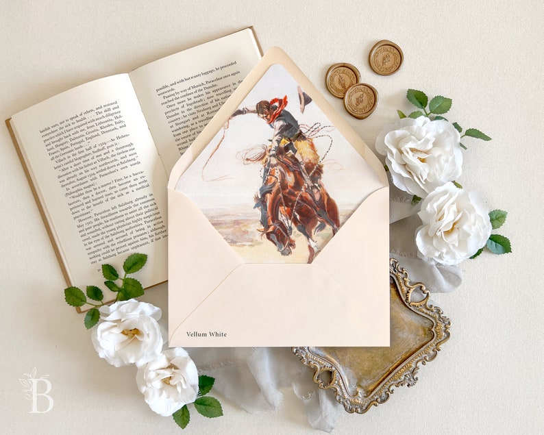 Op de afbeelding: Een cr&egrave;mekleurige envelop met een Vellum White label, voorzien van een binnenvoering met een illustratie van een cowboy op een bokkend paard met een lasso. De envelop ligt naast een open boek, drie gouden waszegels en witte rozen.