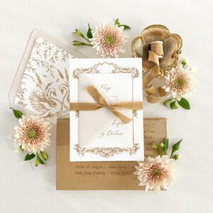 May include: A white wedding invitation with gold calligraphy and a floral design. The invitation is on a brown background with a gold ribbon and flowers.