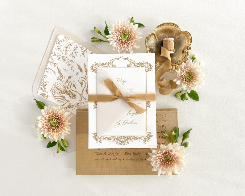 May include: A white wedding invitation with gold calligraphy and a floral design. The invitation is on a brown background with a gold ribbon and flowers.