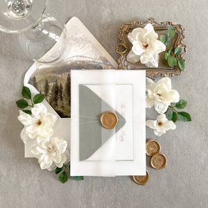 May include: A white envelope with a gray inner envelope and a wax seal. The envelope is tied with a white ribbon and has a floral design on the back. The envelope is surrounded by white flowers and a small gold tray with a white flower.