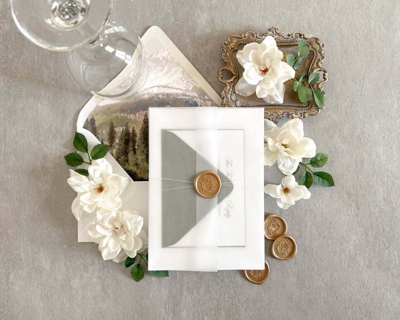 May include: A white envelope with a gray inner envelope and a wax seal. The envelope is tied with a white ribbon and has a floral design on the back. The envelope is surrounded by white flowers and a small gold tray with a white flower.