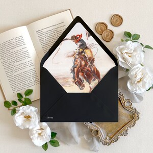 Op de afbeelding: Een zwarte envelop met het woord Ebony op de voorzijde, met een illustratie van een cowboy op een bokkend bruin paard die een lasso vasthoudt. De envelop is omringd door een open boek, witte rozen en gouden waszegels.