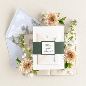 May include: Wedding invitation suite featuring a green and white ribbon, a white envelope, and a white card with the text "Flora and Quinn" in black calligraphy. The invitation is surrounded by blush pink and white flowers.