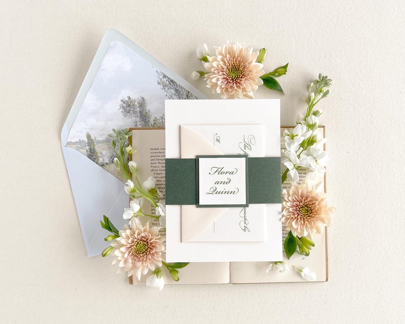 May include: Wedding invitation suite featuring a green and white ribbon, a white envelope, and a white card with the text "Flora and Quinn" in black calligraphy. The invitation is surrounded by blush pink and white flowers.