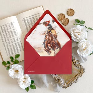 Op de afbeelding: Een dieprode envelop met een puntige klep, voorzien van een binnenvoering bedrukt met een aquarelillustratie van een cowboy op een bokkend paard. Het woord Vermilion is in het wit gedrukt in de linkerbenedenhoek van de envelop.