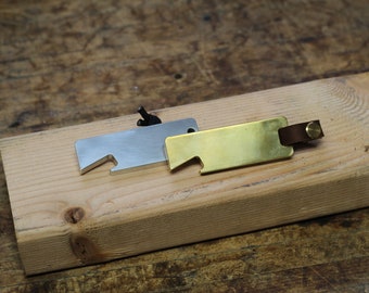 May include: Two bottle openers, one silver and one gold, are shown on a wooden surface. The gold bottle opener has a brown leather strap.