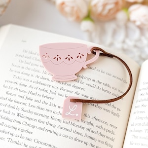 May include: A pink teacup-shaped bookmark with a brown leather cord, resting on an open book. The teacup has decorative cutouts and a small tag with the letter "L". A charming accessory for book lovers.