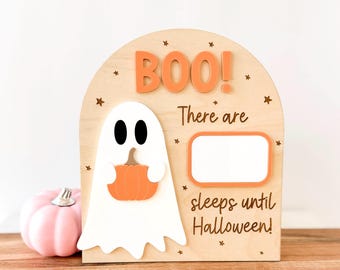 May include: Wooden Halloween countdown sign with a ghost holding a pumpkin. The sign says "BOO!" and "There are sleeps until Halloween!" in orange lettering. A pink pumpkin sits to the left of the sign.