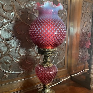 May include: A vintage red hobnail glass lamp with a ruffled white top. The lamp has a brass base and a heart-shaped midsection. The lamp is on a wooden surface with ornate carved details in the background.