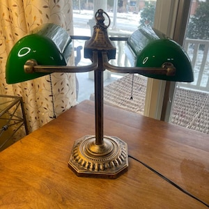 May include: A double-headed banker lamp featuring two emerald green glass shades and a dark bronze-toned metal base with an octagonal pedestal. The lamp includes two adjustable arms and two pull-chain switches hanging from each shade.