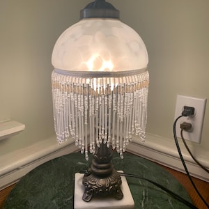 May include: A table lamp with a frosted white glass dome shade and a decorative fringe of clear and cream-colored glass beads. The lamp features an ornate, dark metallic base with a square white marble plinth and a small finial at the top.