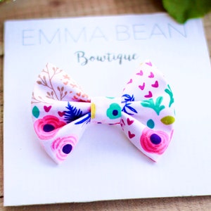 May include: A pink fabric bow with a floral and heart print. The bow is on a white card with the text "Emma Bean Boutique".
