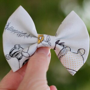 May include: A white fabric bow with a black and white print of cartoon animals and yellow flowers. The bow is tied in the center with a yellow flower.