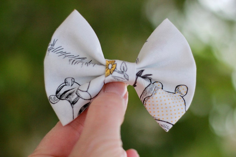 May include: A white fabric bow with a black and white print of cartoon animals and yellow flowers. The bow is tied in the center with a yellow flower.