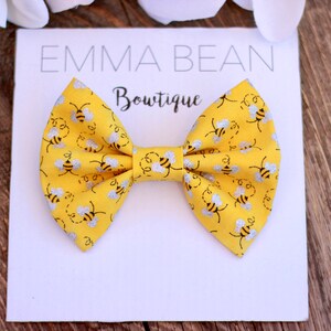 May include: A yellow bow with a silver glitter bee pattern. The bow is on a white card with the text "Emma Bean Bowtique".