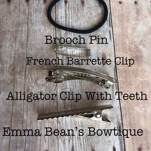 May include: A close-up of four different hair accessories on a wooden surface. The accessories include a ponytail band, a brooch pin, a French barrette clip, and an alligator clip with teeth. The text "Emma Bean's Bowtique" is visible at the bottom of the image.