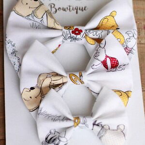 May include: Three white fabric bow ties with a Winnie the Pooh and friends print. The bow ties feature yellow, red, and brown accents. The bow ties are arranged in a stack, with the largest bow tie on the bottom and the smallest bow tie on top.