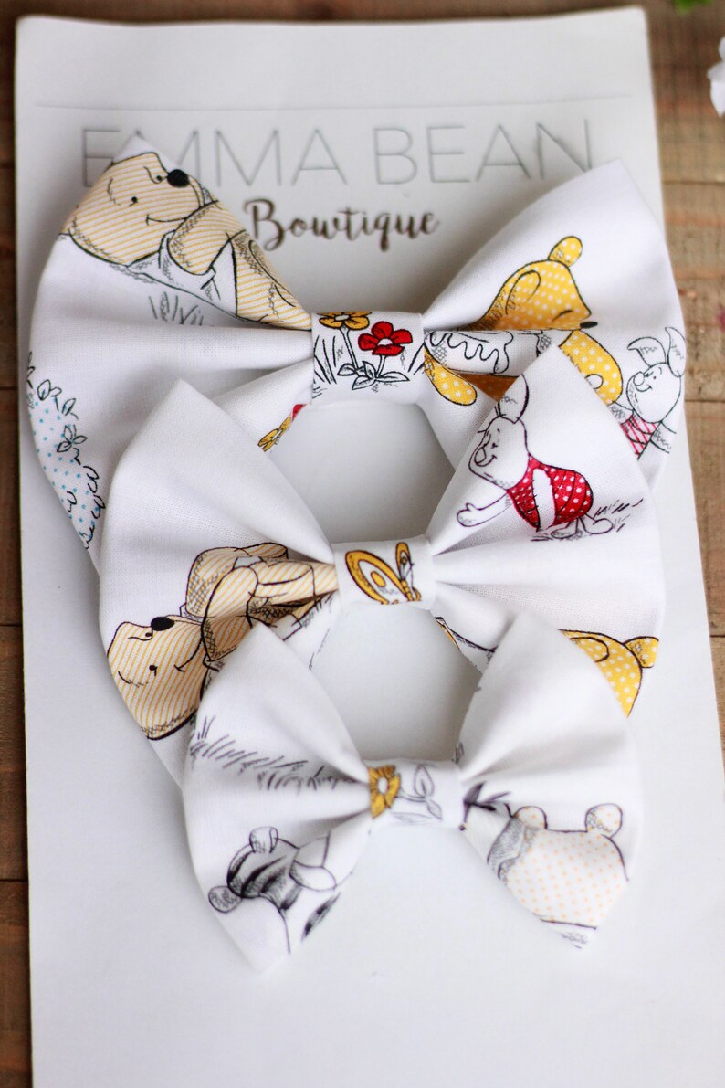 May include: Three white fabric bow ties with a Winnie the Pooh and friends print. The bow ties feature yellow, red, and brown accents. The bow ties are arranged in a stack, with the largest bow tie on the bottom and the smallest bow tie on top.